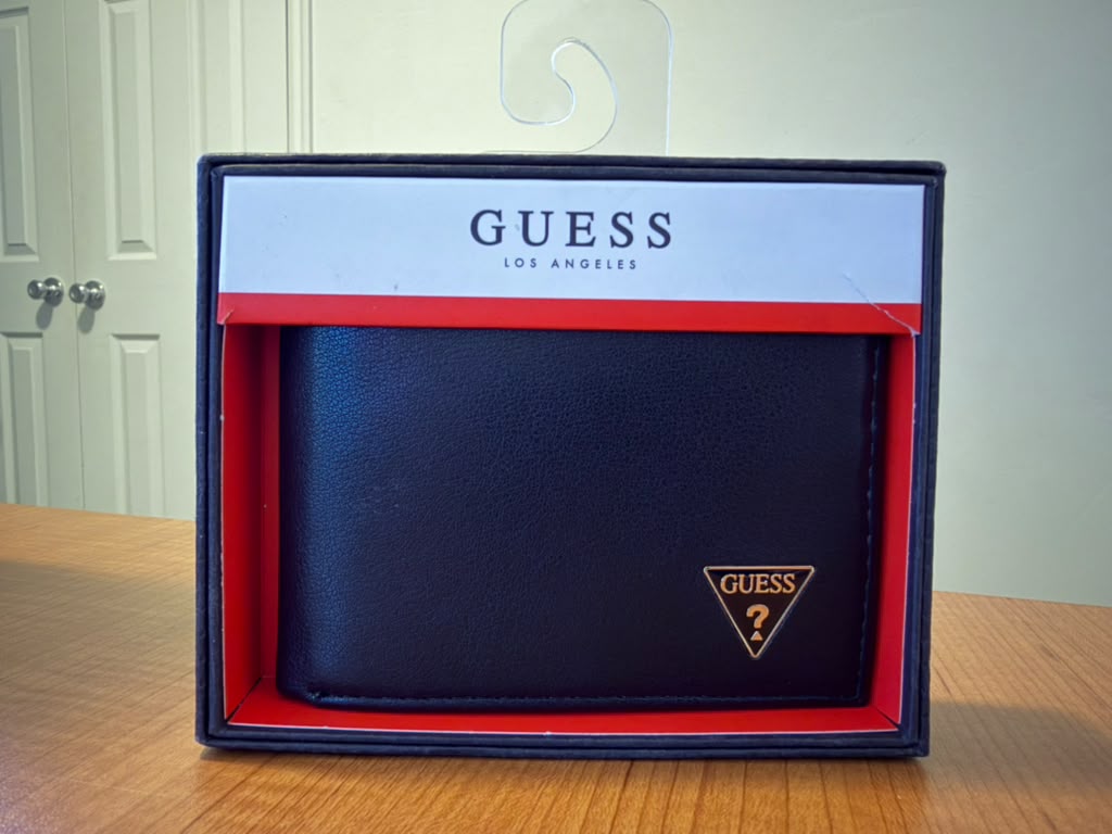 Billetera Guess color negro de acabado liso, diseño plegable, múltiples compartimentos para tarjetas y billetes, detalle frontal con logo triangular Guess, presentación original en caja, ideal para uso diario o regalo.