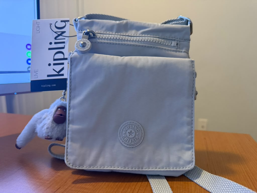 Bolso Kipling color gris claro, diseño ligero con múltiples compartimentos y bolsillos con cierre, correa ajustable para usar al hombro o cruzada, incluye el clásico llavero de mono Kipling, ideal para uso diario, viajes o llevar artículos esenciales, nuevo con etiqueta.