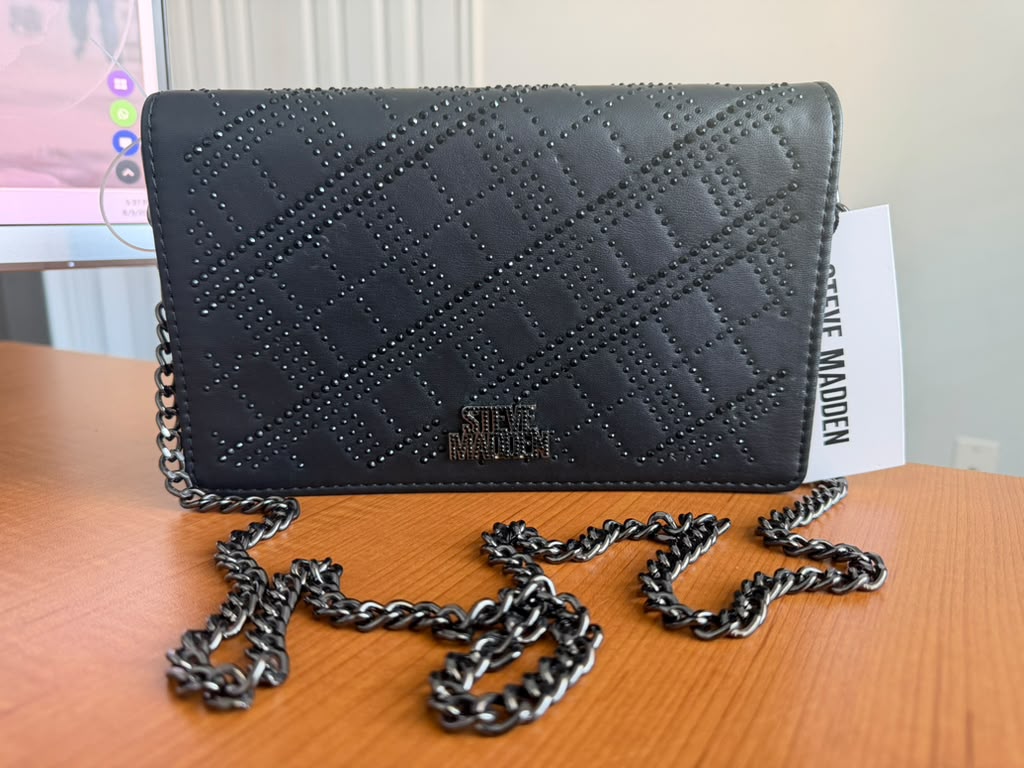 Steve Madden. Cartera tipo crossbody color negro con diseño texturizado, cierre superior, solapa frontal, logo metálico y correa de cadena.