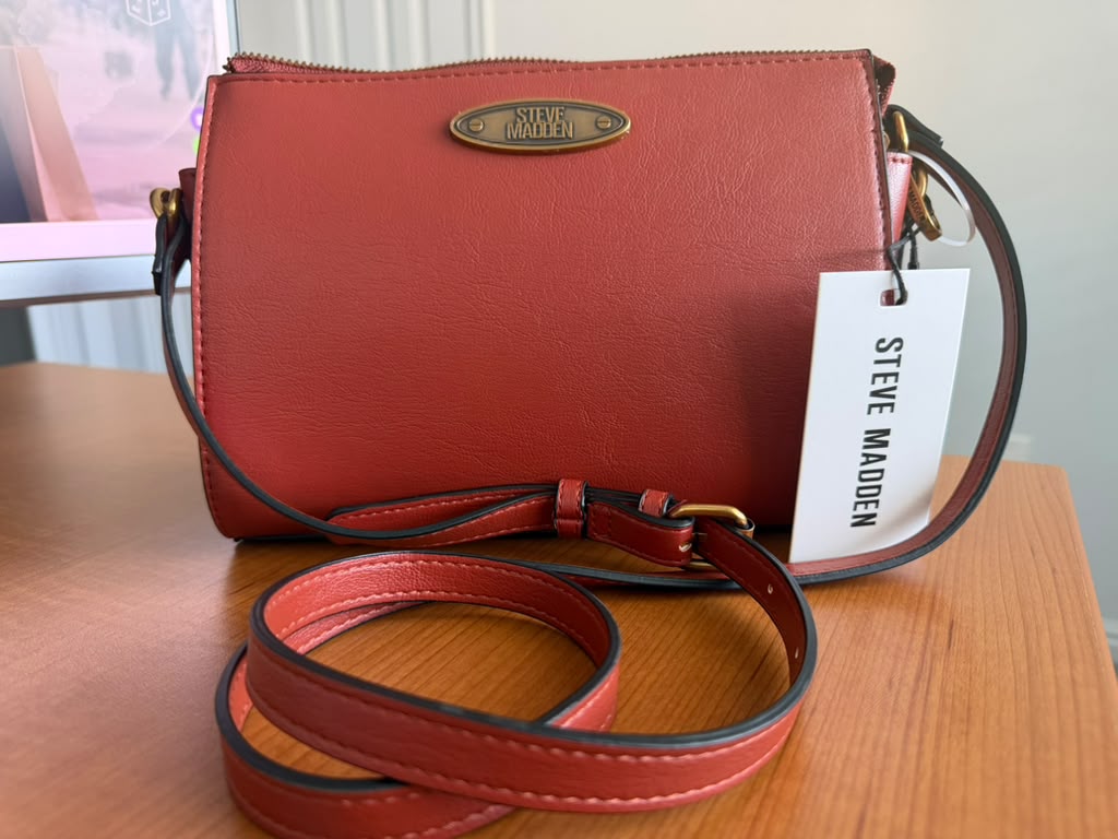 Steve Madden. Cartera crossbody color terracota, diseño liso, cierre superior, correa ajustable y removible, logo metálico frontal.