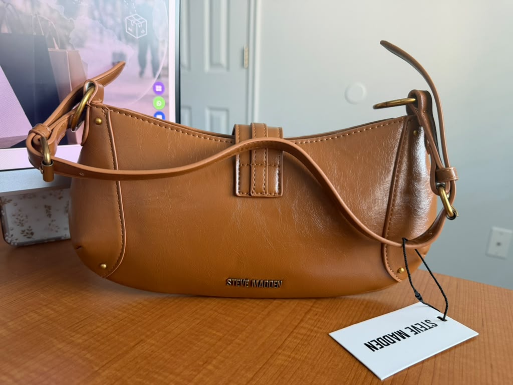 Steve Madden. Cartera de hombro color camel, diseño liso, cierre superior, correa ajustable, herrajes dorados y logo metálico frontal.