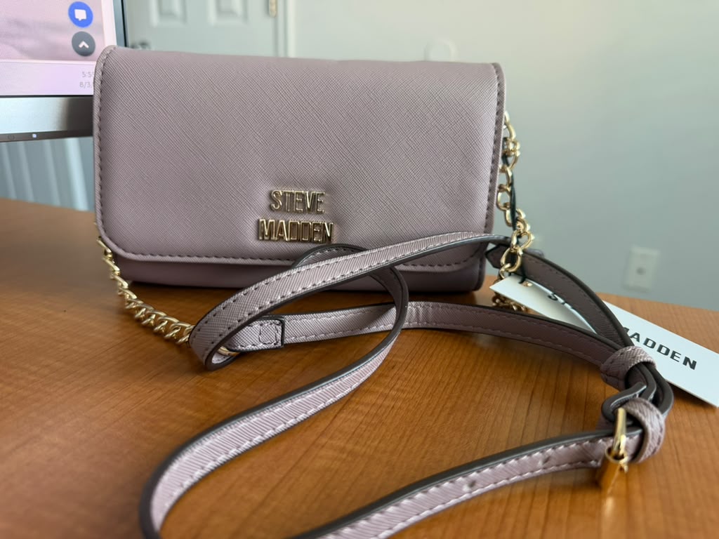 Steve Madden. Cartera crossbody color lavanda, diseño texturizado, solapa frontal, correa ajustable con cadena y logo metálico.