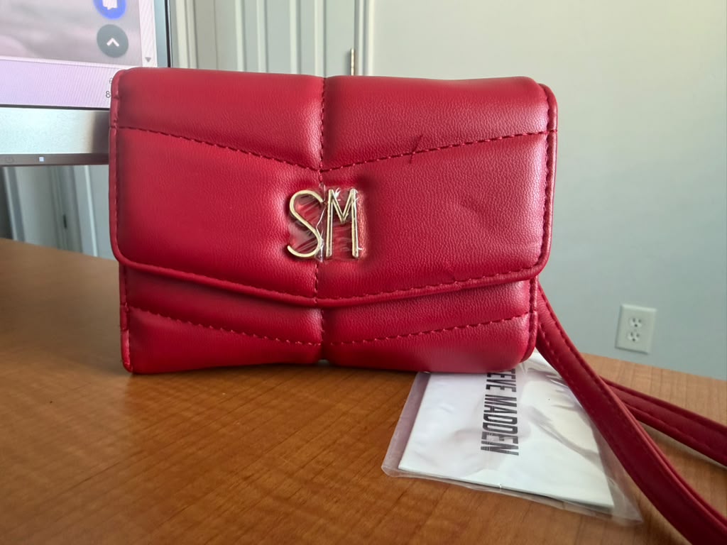 Steve Madden. Billetera acolchada color rojo, diseño moderno con broche frontal, logo metálico SM y correa de muñeca desmontable.